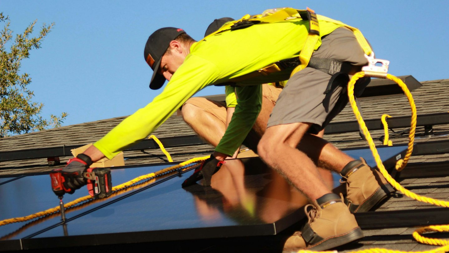 Hombre instala paneles solares en el techo de una casa con un taladro.